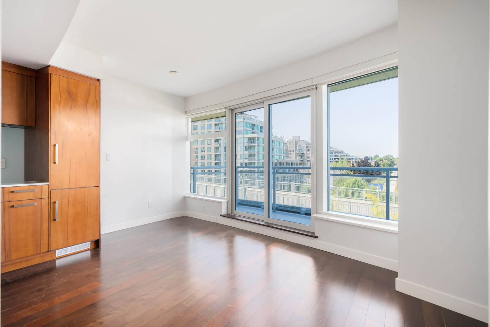 Sunlit, empty room with large windows, hardwood floors, and balcony door facing modern buildings on 1565 W 6th Ave, Vancouver.