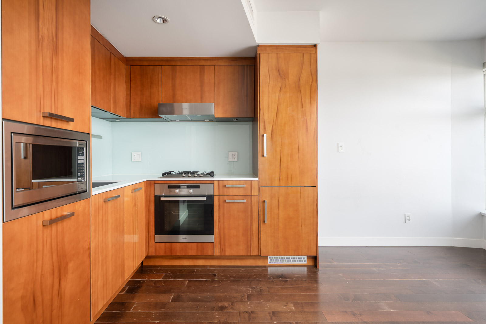 Modern kitchen in Vancouver with wooden cabinets, built-in oven, microwave, gas stove, glass backsplash, and dark floor.