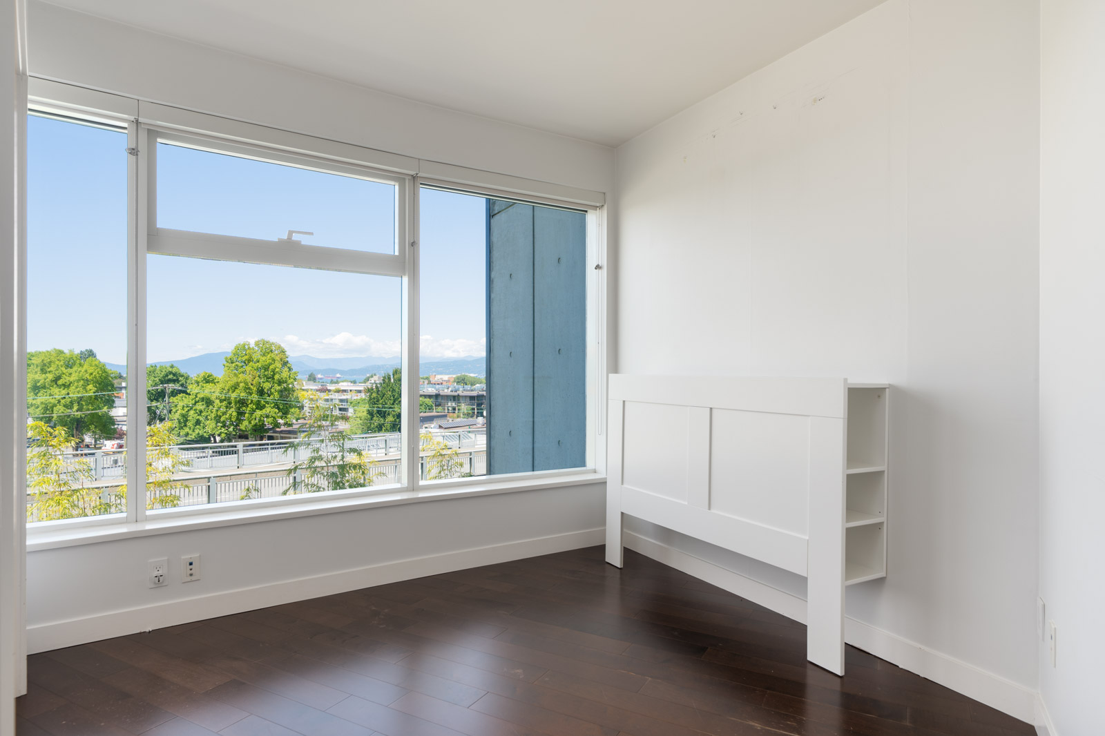 Empty room with dark wood floor, white walls, large windows showing Vancouver trees and mountains, and a white bookshelf headboard.