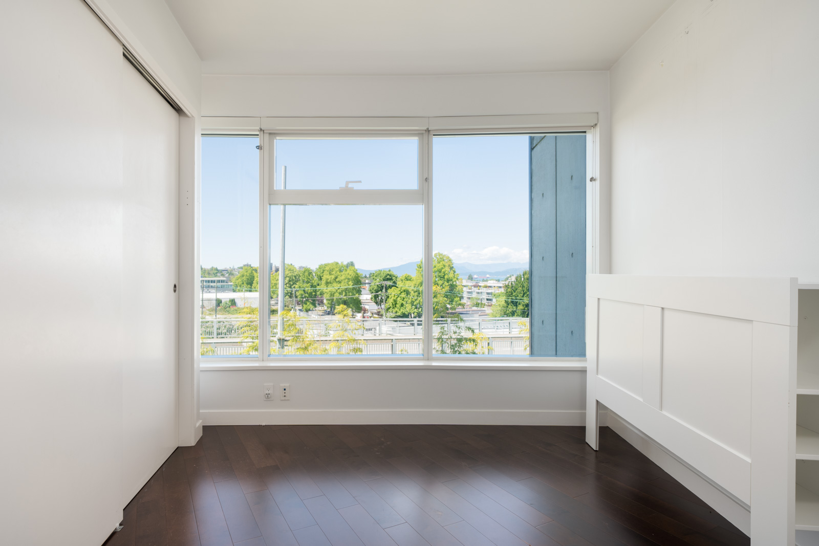 Empty room with dark wood floors, white walls, sliding closet door, and large windows overlooking trees and buildings in Vancouver.