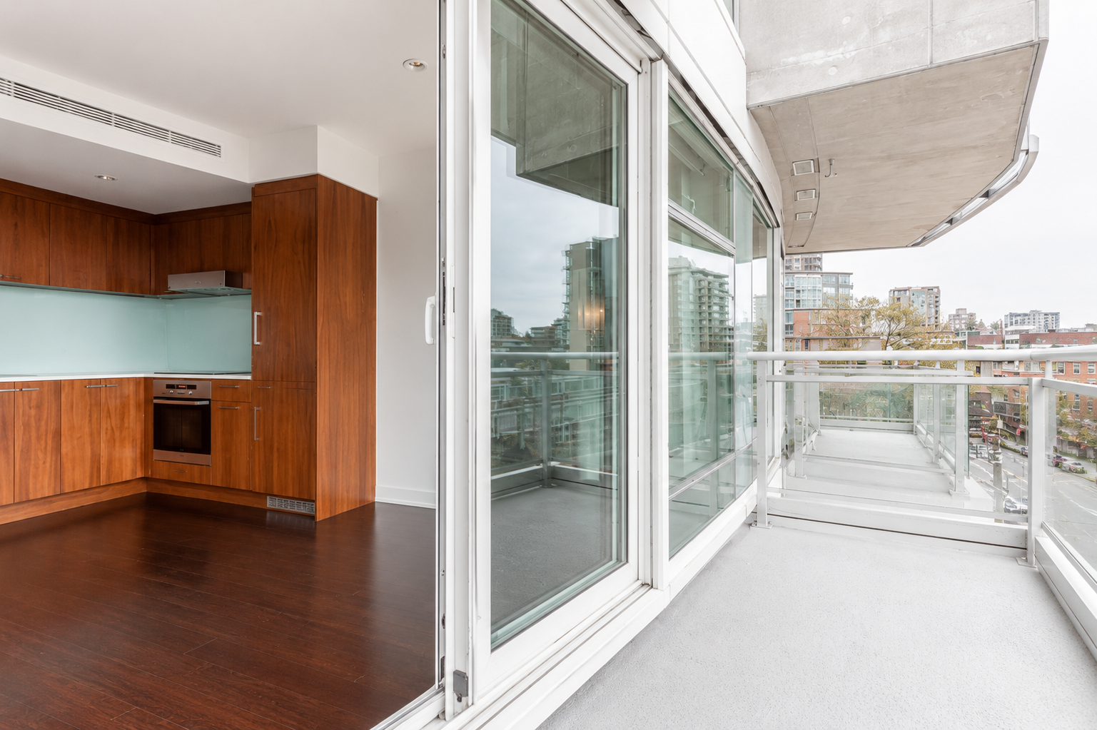 Modern apartment with wood kitchen, dark floors, and large glass doors opening to a spacious balcony overlooking an urban cityscape.