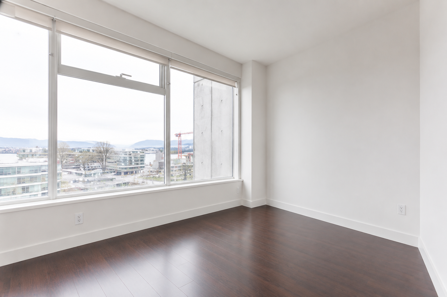 Empty room with white walls, dark wood flooring, and large windows overlooking a cityscape with buildings and distant mountains.