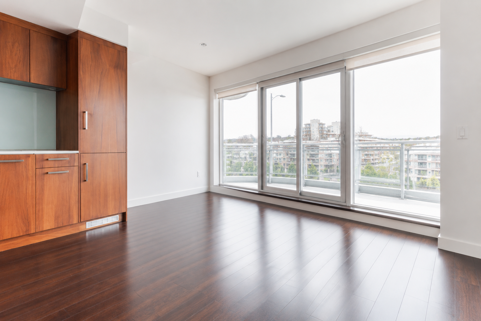 Empty modern apartment with large glass doors leading to a balcony, wood cabinets, and dark hardwood floors, overlooking a cityscape.