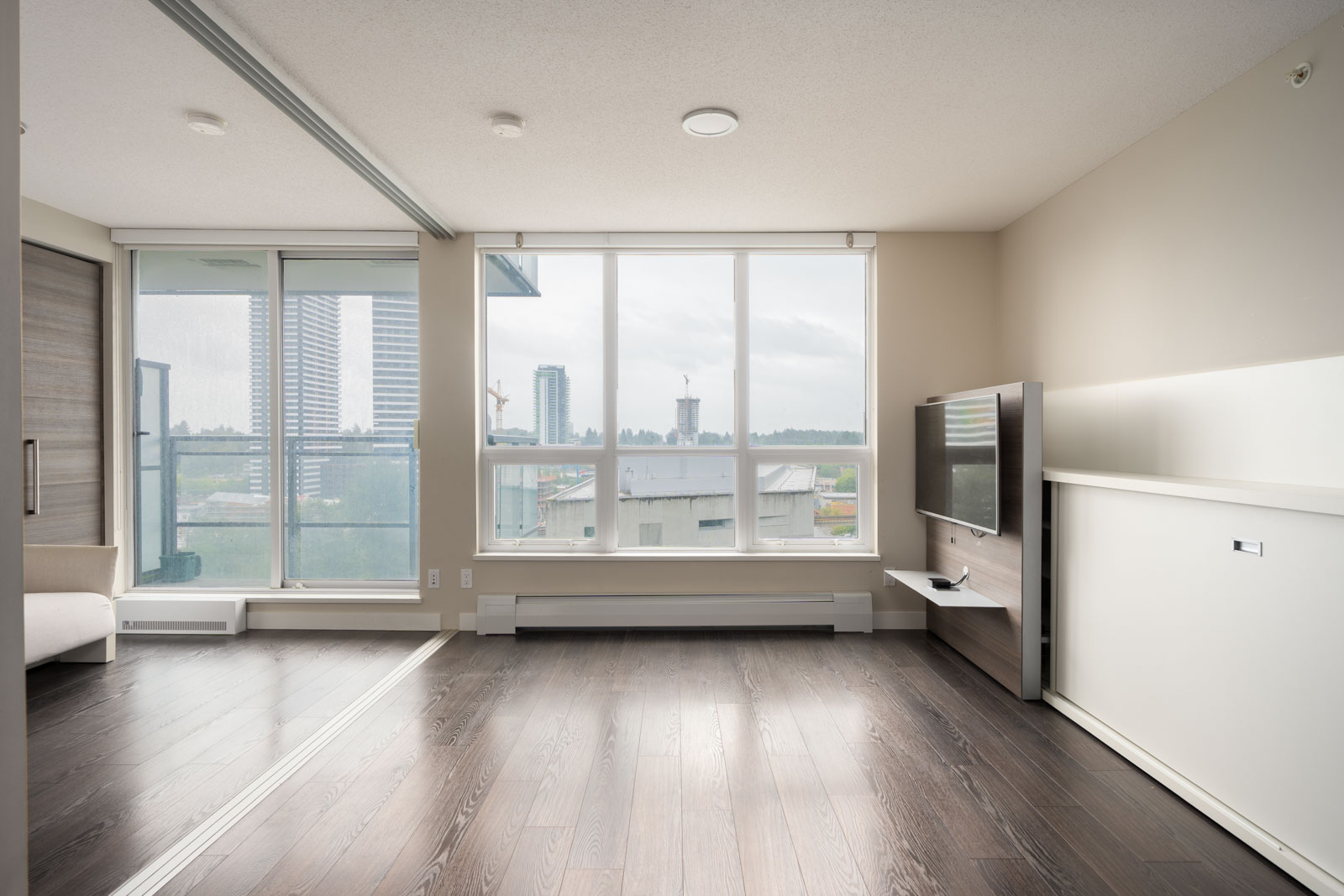Unfurnished modern apartment living room with large windows, dark wood floors, a wall-mounted TV, and a view of city buildings and a balcony outside.