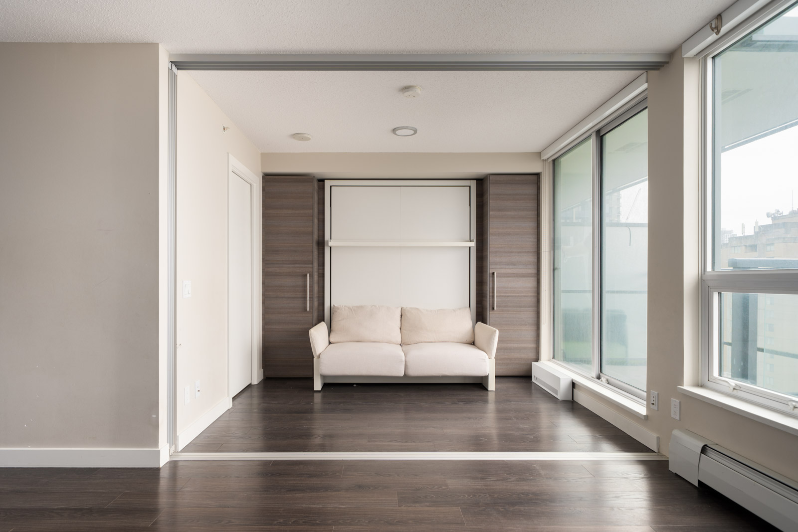 A modern, unfurnished room with dark wood floors, large windows, and a beige sofa positioned in front of a Murphy bed housed in a built-in wooden unit.