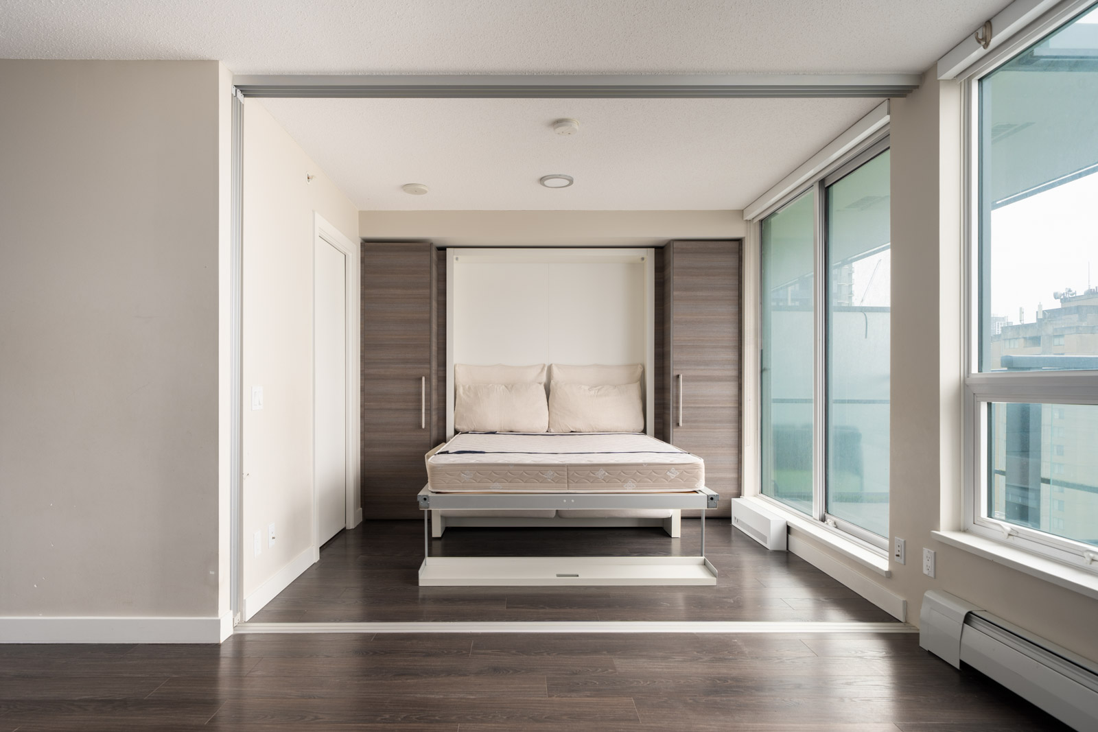 A modern studio apartment with a Murphy bed partially pulled down, wood floors, large windows, and neutral-colored walls.