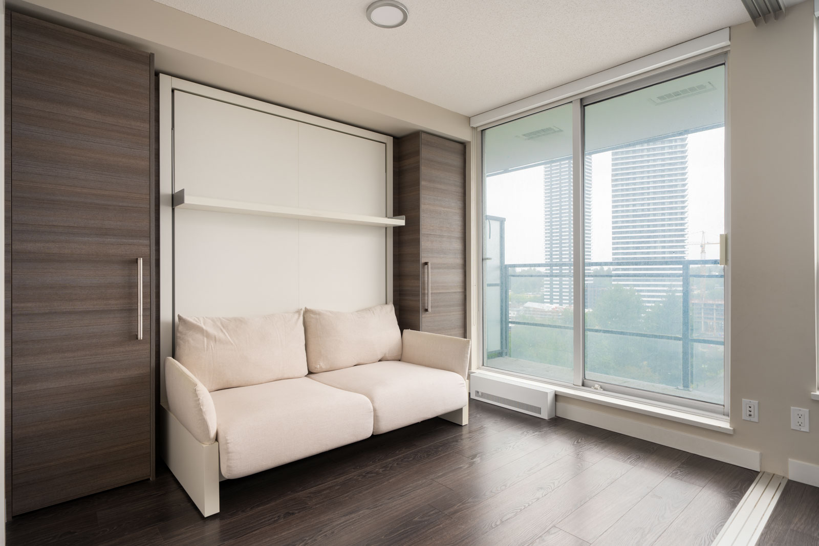 A modern room with a beige sofa in front of a Murphy bed, dark wood floors, large windows, and a city view.