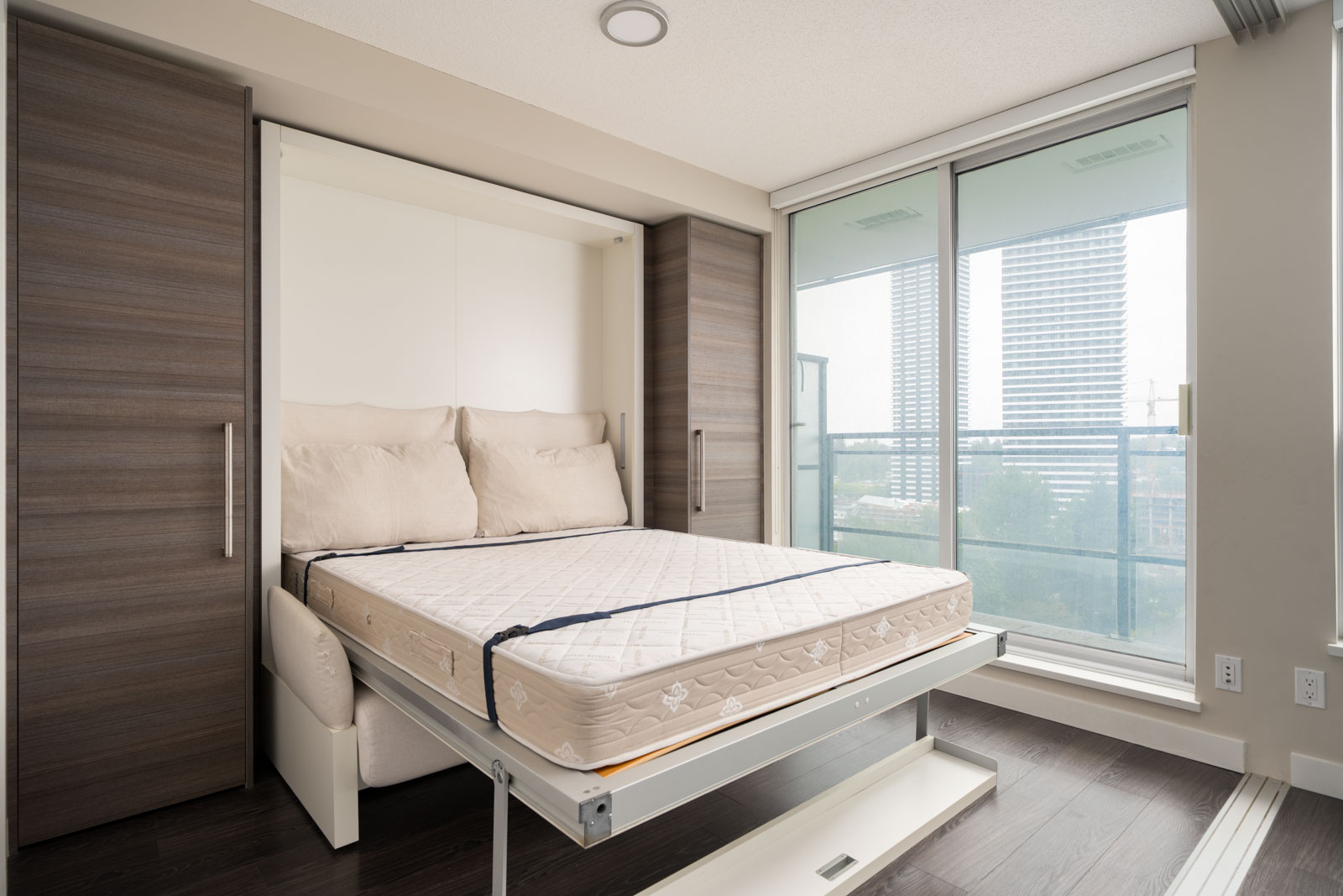 A modern bedroom features a Murphy bed with beige bedding, flanked by two wardrobes, and large windows offering a city view.