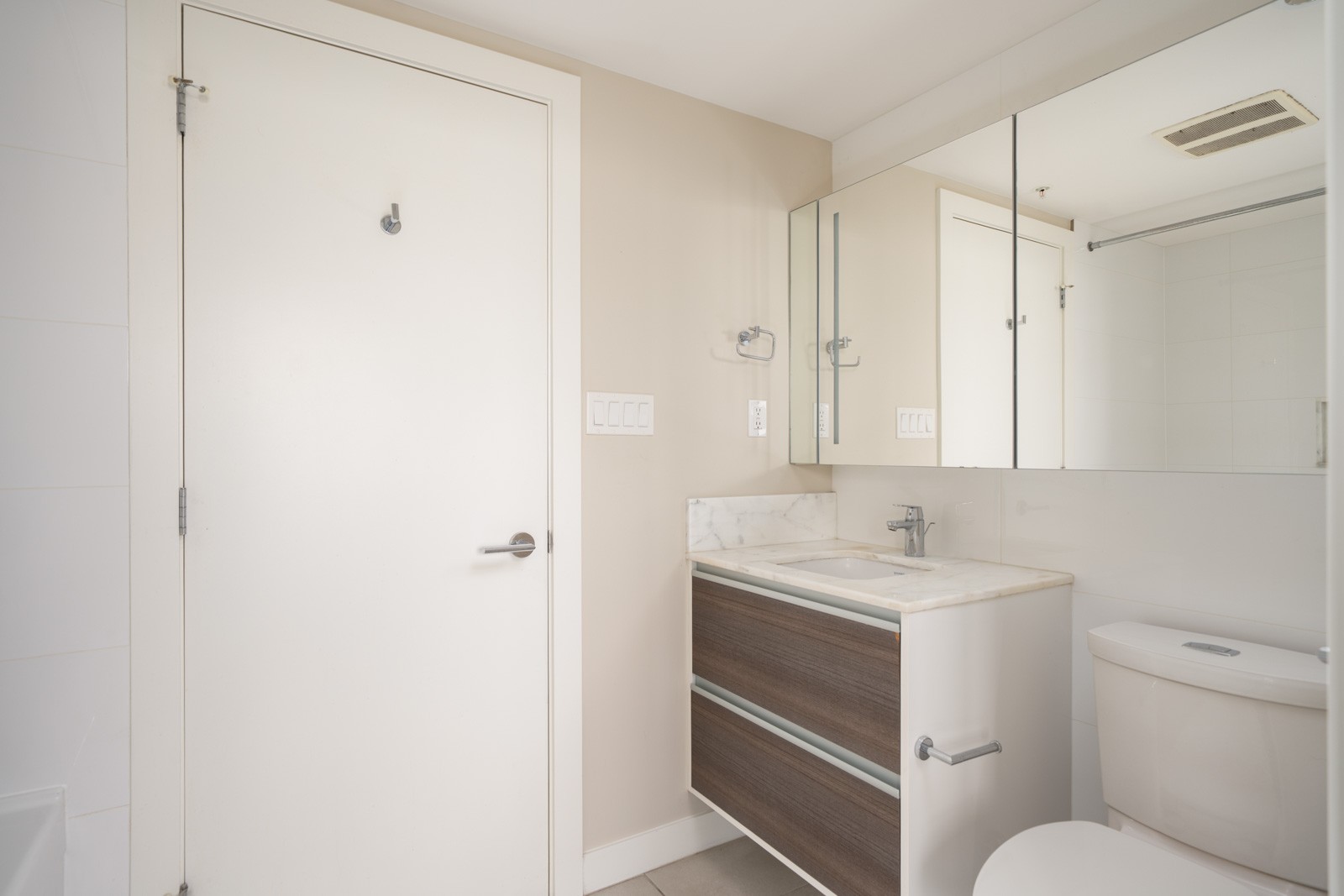 Minimalist bathroom with a white door, a modern vanity with a marble countertop, a wall-mounted mirror cabinet, and a toilet. Neutral color scheme with clean lines.