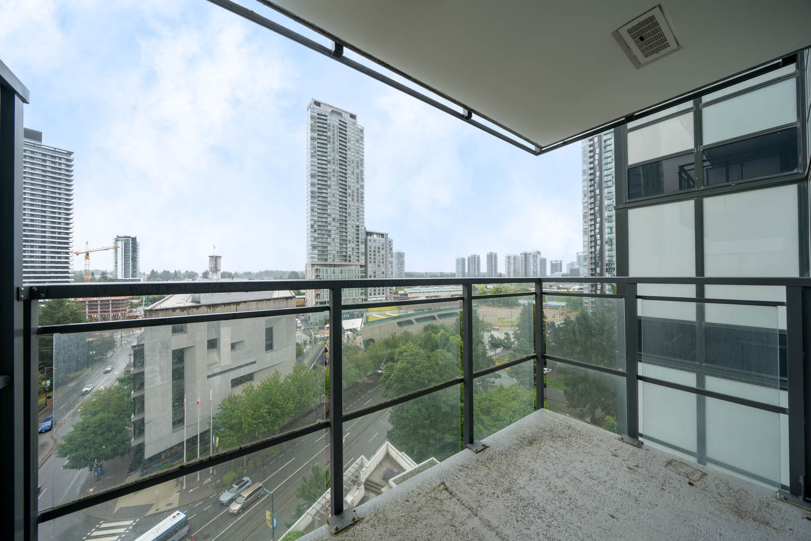 View from a high-rise balcony overlooking a cityscape with tall buildings, busy roads, and trees on a clear day.