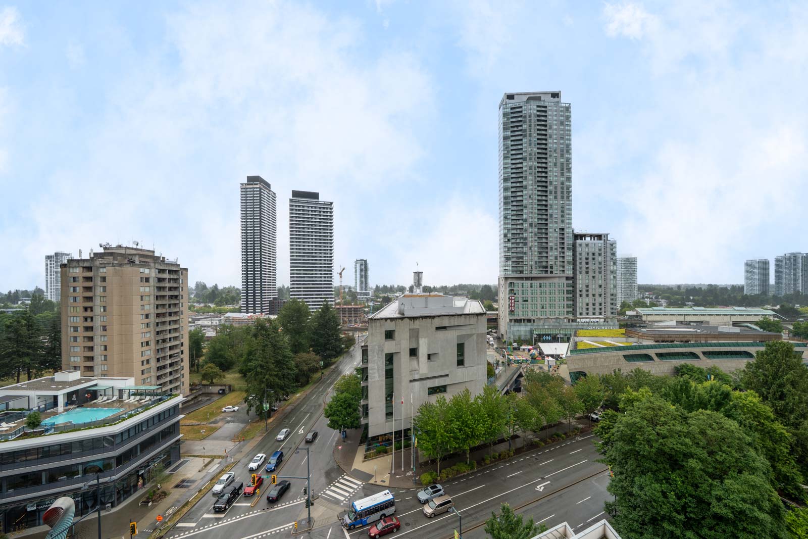 A cityscape with several high-rise buildings, roads with cars, a mix of modern and older structures, and trees lining the streets under a partly cloudy sky.