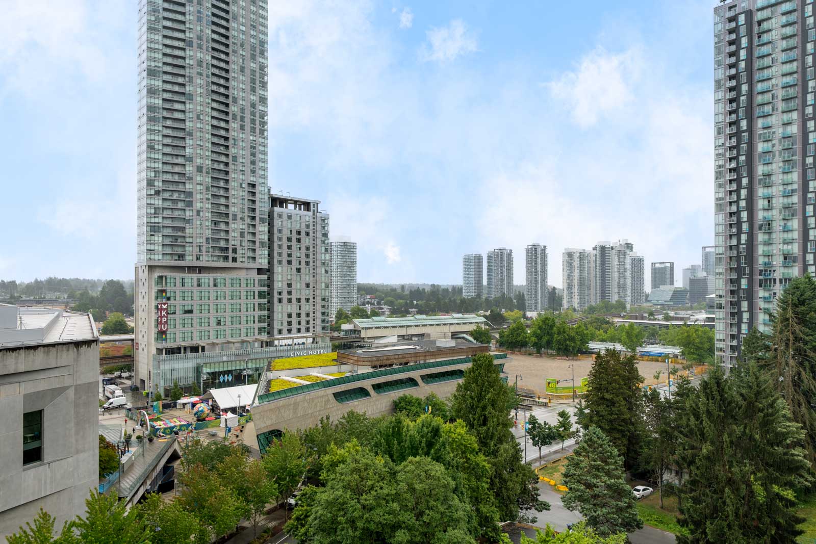 A cityscape with tall modern buildings, green trees, and a construction site in the foreground under a partly cloudy sky.