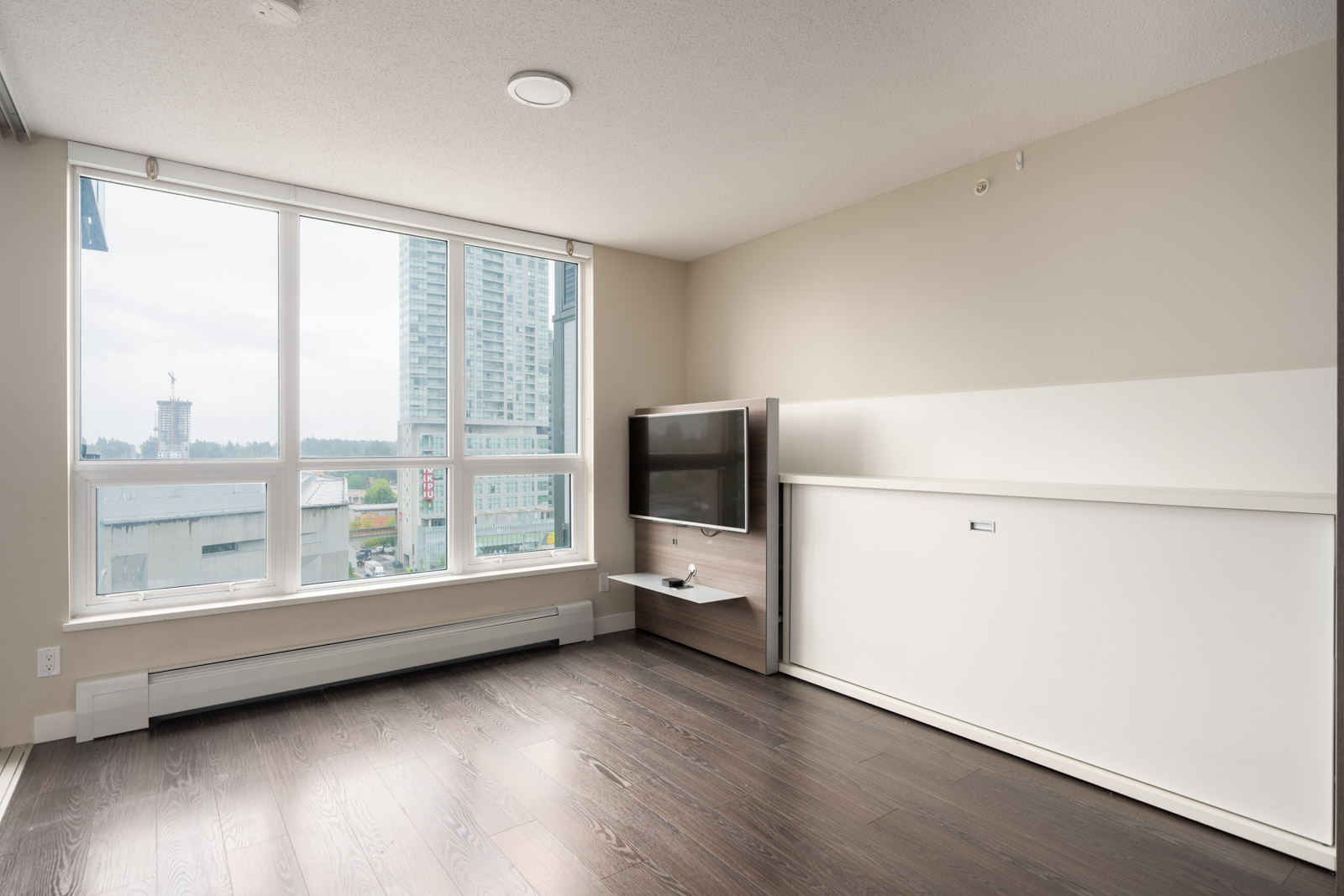 Unfurnished apartment room with large window, wall-mounted TV, hardwood floors, and a closed Murphy bed against a beige wall. City buildings are visible outside the window.