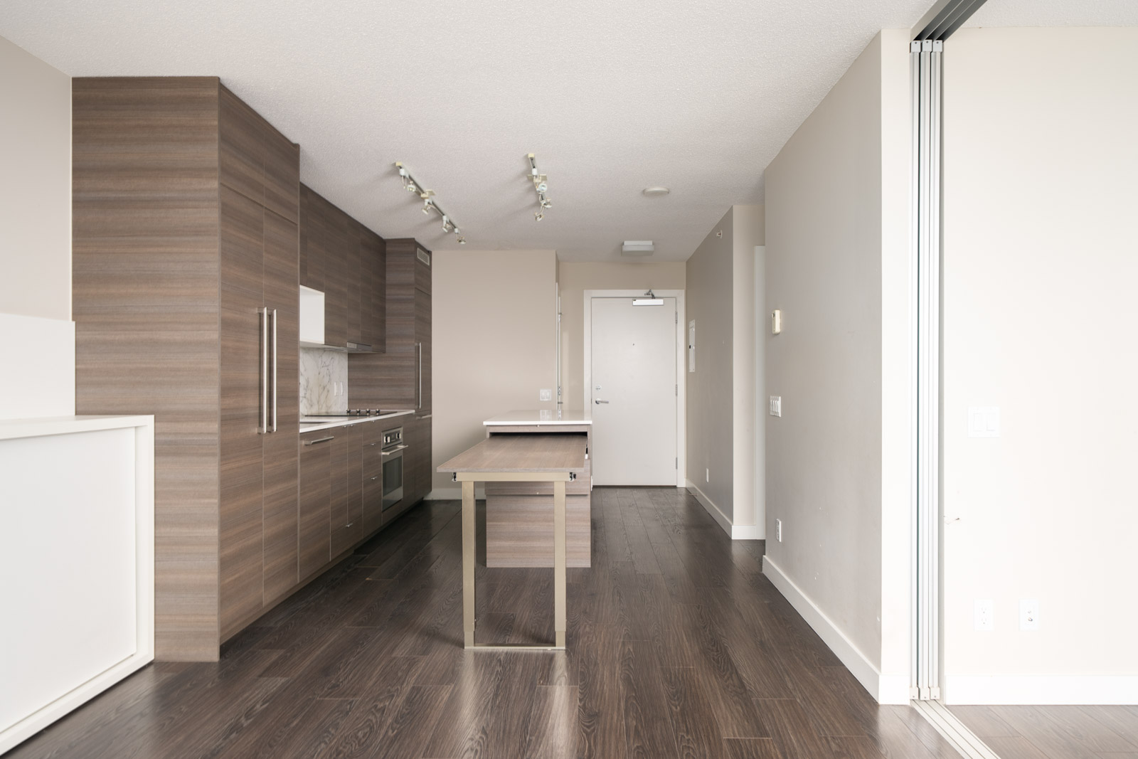 Modern apartment kitchen with wood cabinets, a small island, dark hardwood floors, and beige walls, viewed from the living area with ample natural light.