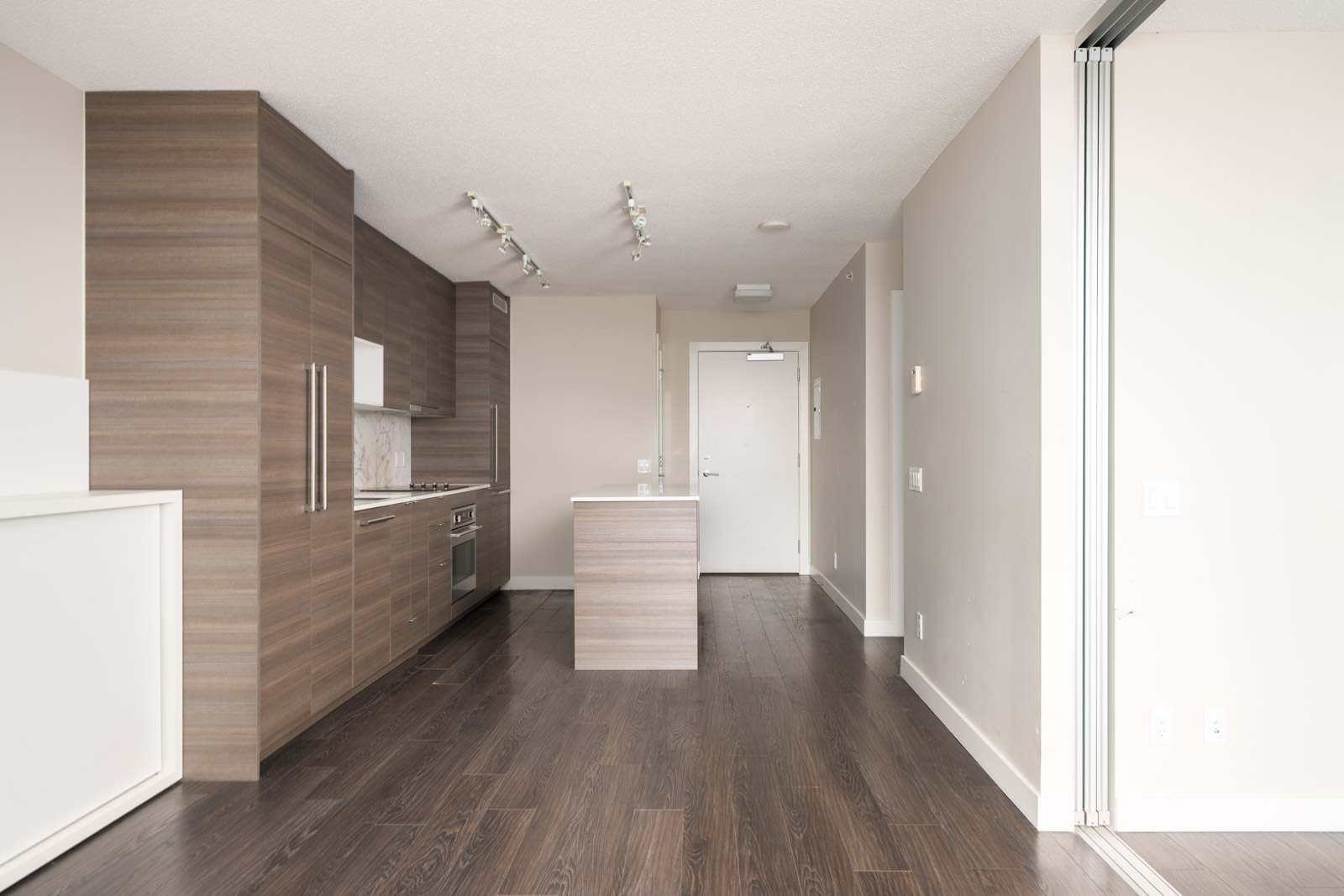 Modern apartment kitchen with wood cabinetry, dark hardwood floors, recessed lighting, and neutral-colored walls, viewed from an empty living area.