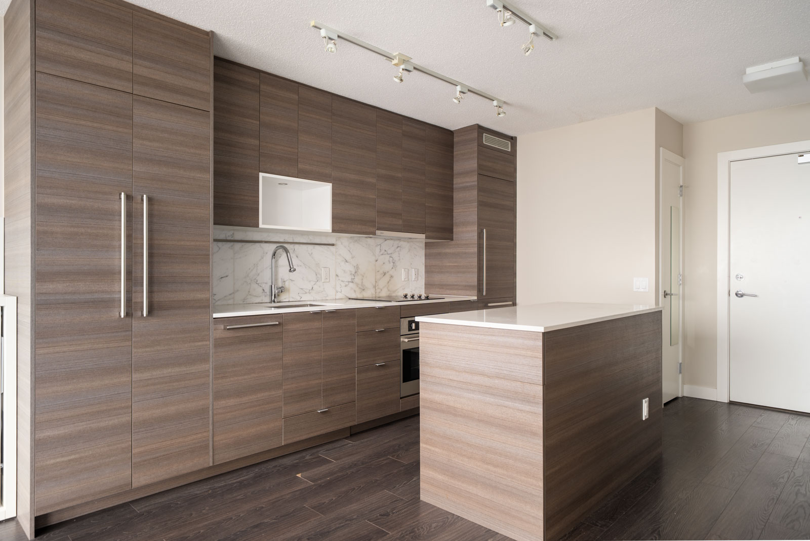 Modern kitchen with wood cabinetry, marble backsplash, built-in oven, electric cooktop, and a peninsula countertop, featuring neutral tones and dark wood flooring.