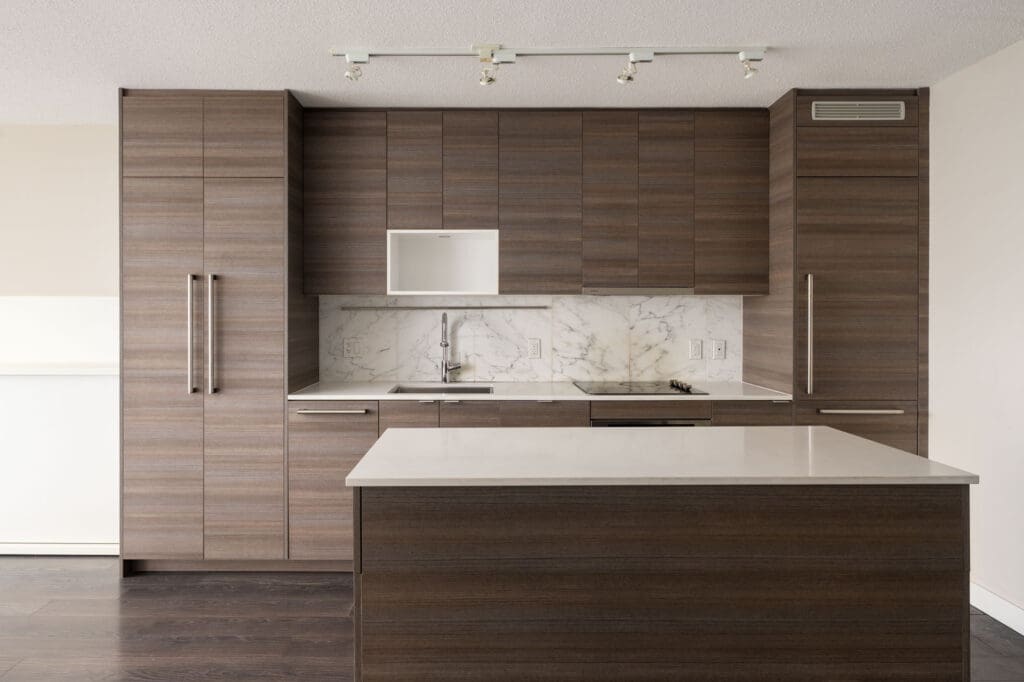 Modern kitchen with wood cabinetry, white marble backsplash, built-in appliances, and a white countertop island under ceiling track lighting.