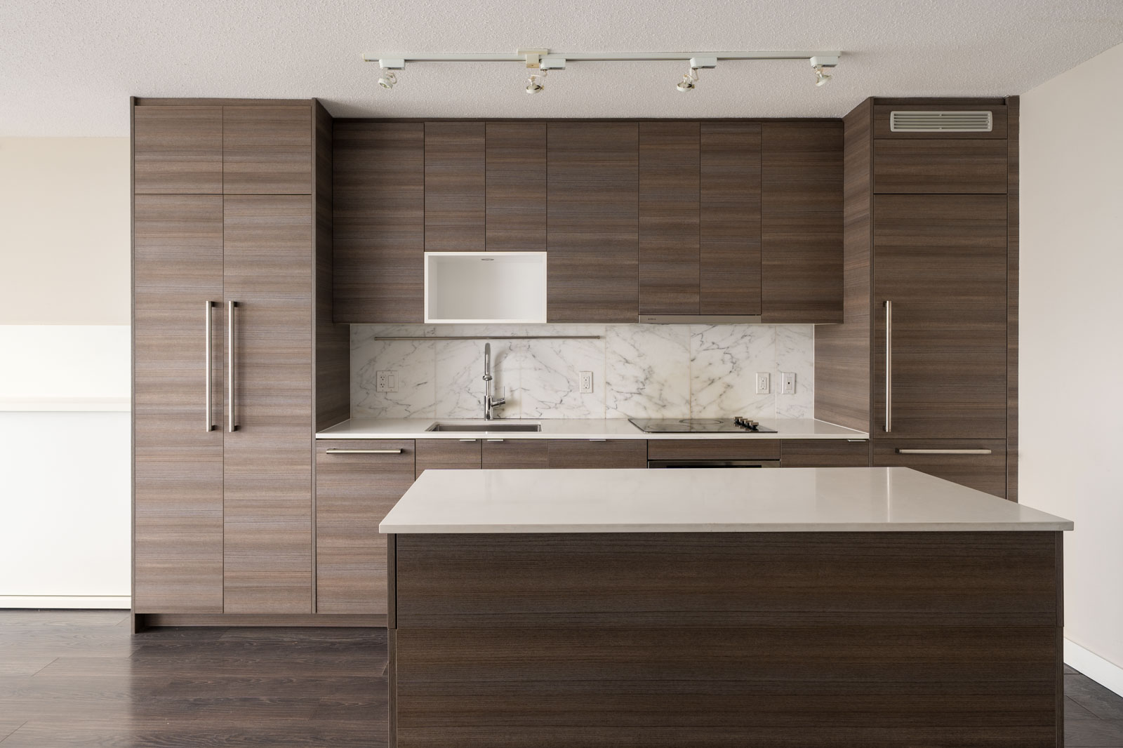 Modern kitchen with wood cabinetry, white marble backsplash, built-in appliances, and a white countertop island under ceiling track lighting.