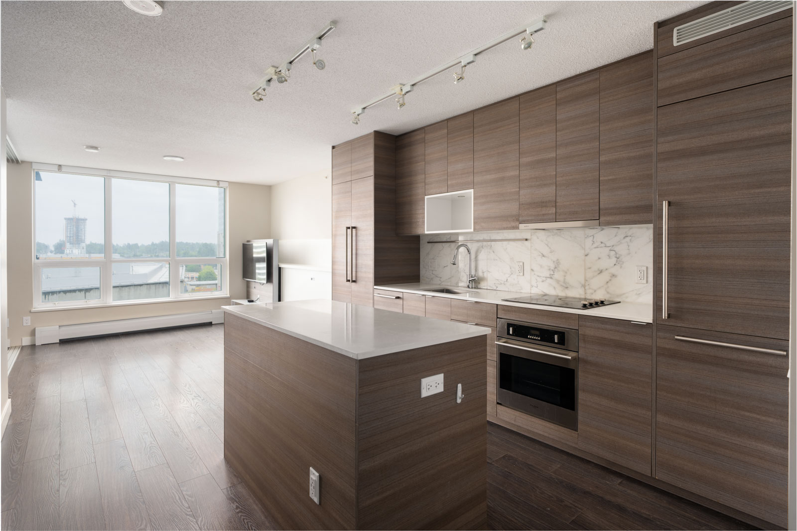 Modern kitchen with wood cabinets, marble backsplash, built-in appliances, and a central island in an open-plan apartment with large windows and dark wood flooring.