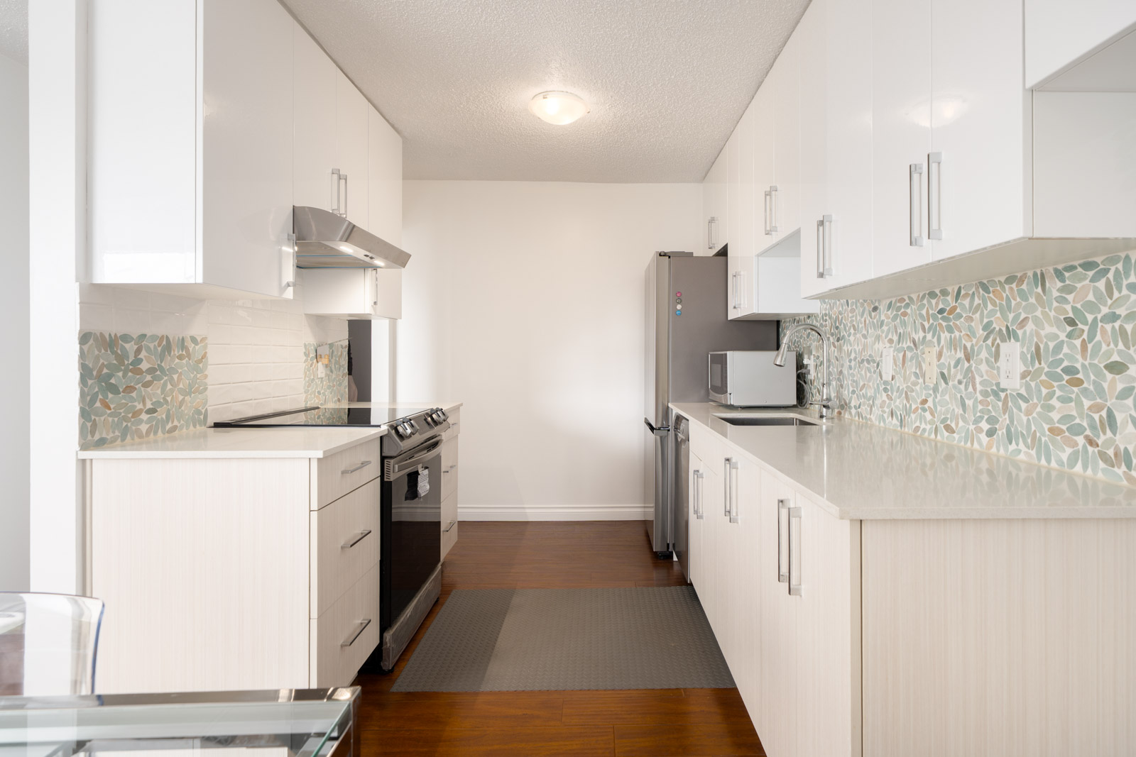 Modern kitchen with white cabinets, stainless steel appliances, gray fridge, mosaic tile backsplash, wood floors, light counters.
