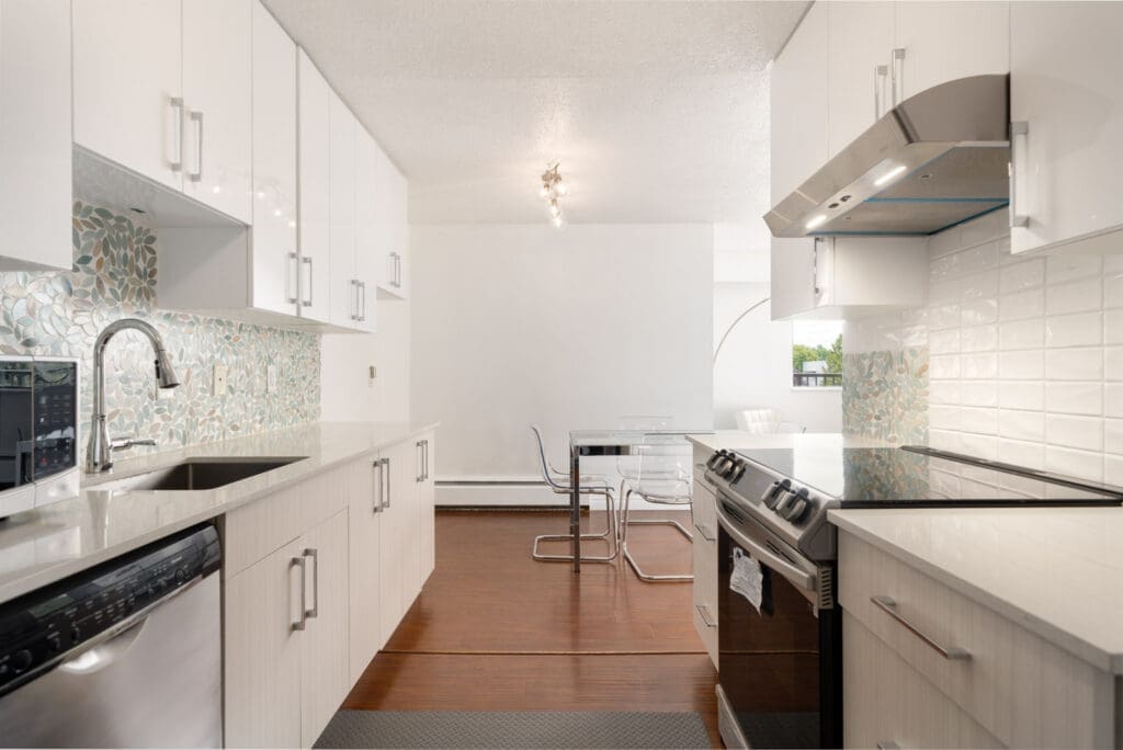 Modern kitchen with white cabinets, stainless steel appliances, glass tile backsplash, and dining area with clear table on wood floor.