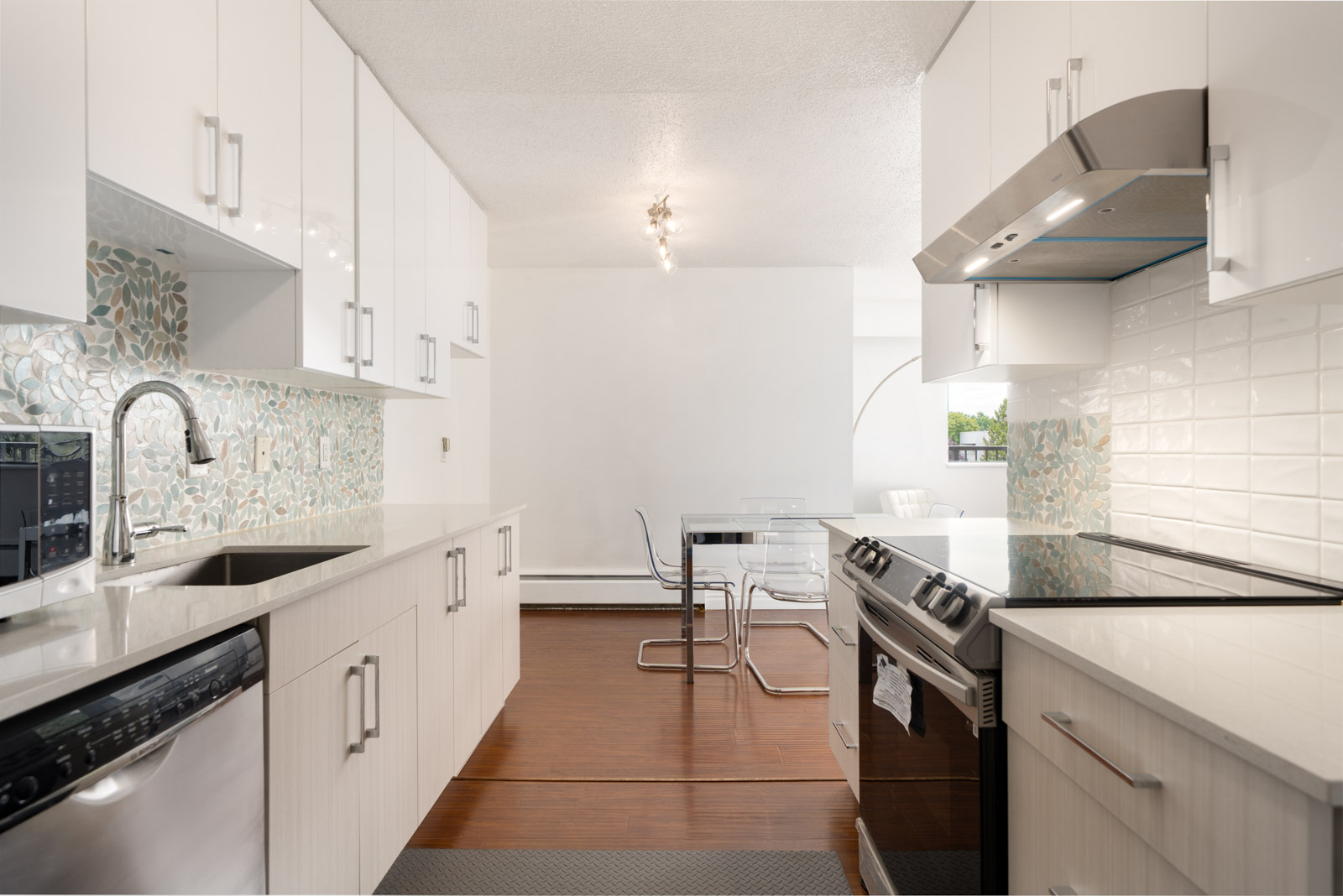 Modern kitchen with white cabinets, stainless steel appliances, glass tile backsplash, and dining area with clear table on wood floor.