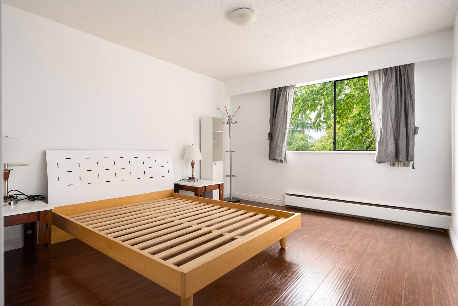 Minimalist bedroom featuring a wooden bed, two side tables with lamps, a coat rack, and a window with gray curtains and tree views.