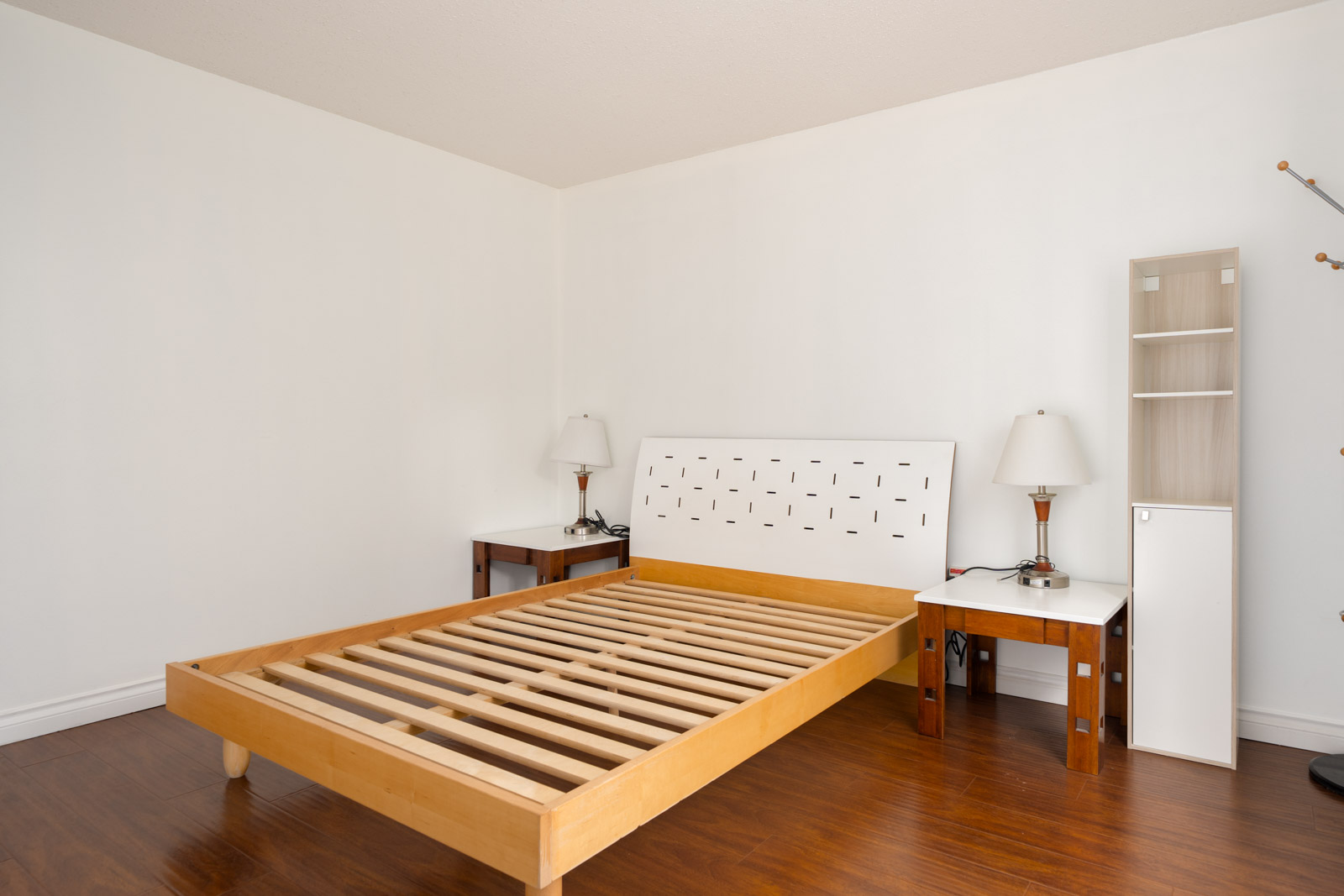 Minimalist bedroom featuring an empty wooden bed frame, white headboard, side tables with lamps, shelving, and wood floors.