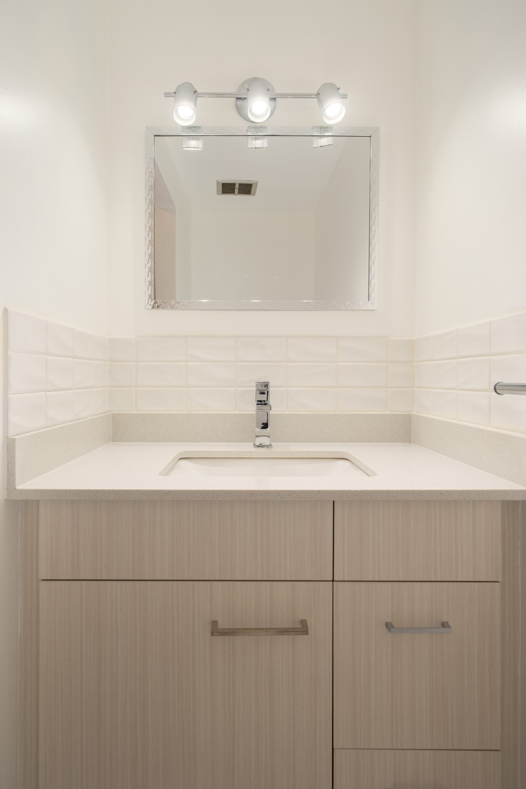 Modern bathroom vanity with a rectangular sink, light wood cabinets, wall-mounted faucet, mirror, and overhead lighting. White tiled walls.