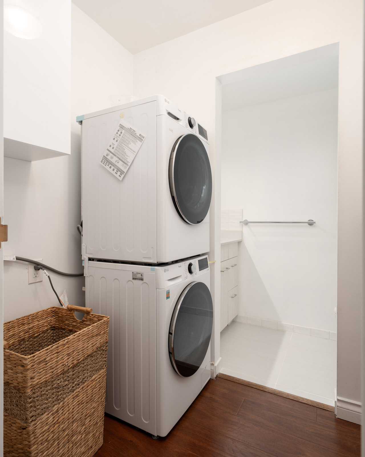 Stacked washer and dryer beside a woven laundry basket on wood floor; open doorway to white tiled room with towel bar.