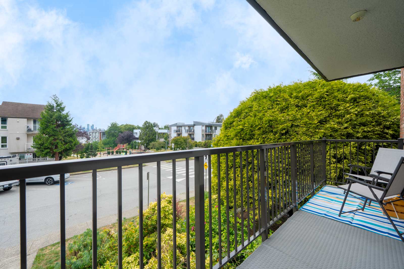 Second-floor balcony with two chairs and a blue-striped rug overlooks street, trees, and homes on a clear day.