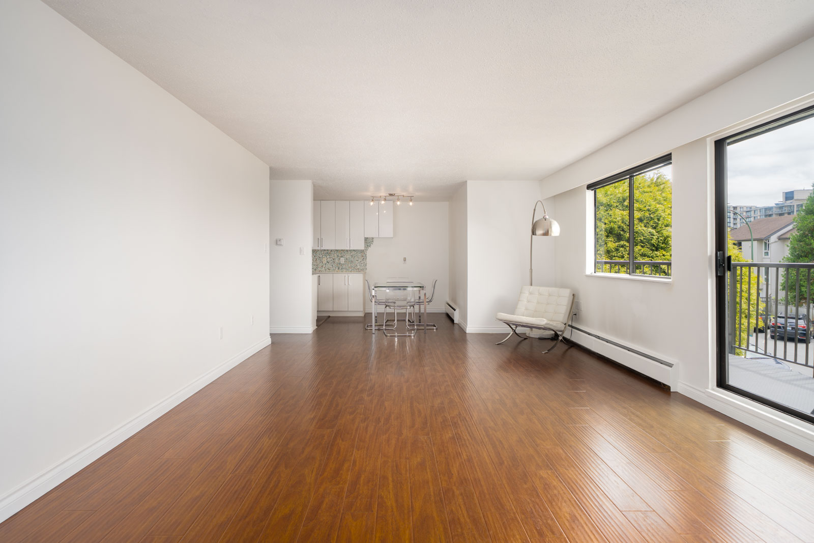 Unfurnished apartment with wood floors, white walls, small dining area, large windows, and glass door to balcony overlooking city.