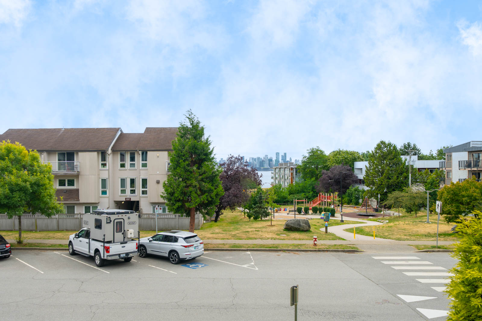 Apartment buildings and parked cars line a grassy park with a playground, trees, and distant city skyline under a blue, partly cloudy sky.