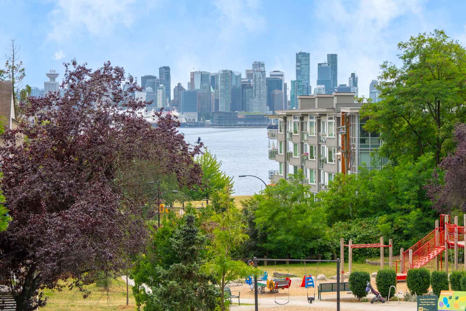 City skyline seen across the water with modern apartments, trees, and a playground visible in the foreground.