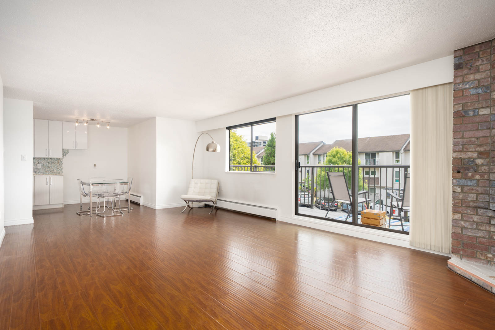 Airy living room with wood floors, brick accent wall, large windows, and sliding door to balcony; white kitchen and dining area behind.