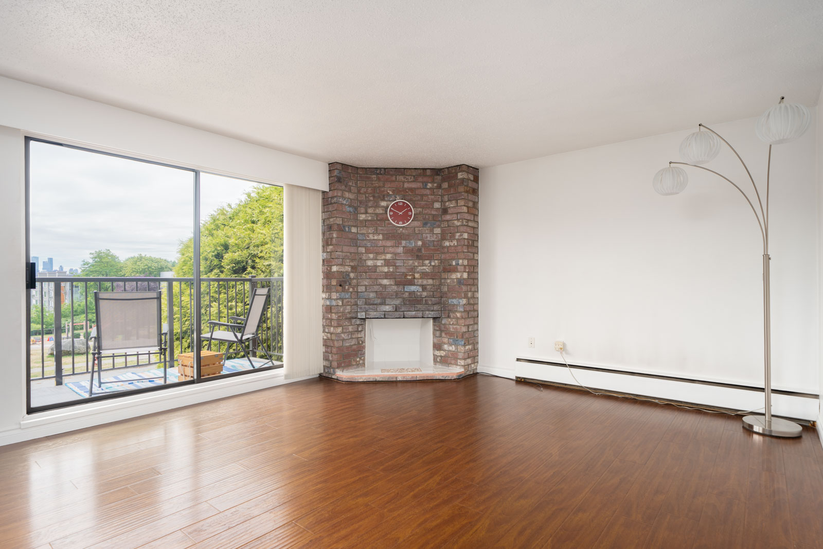 Unfurnished living room with wood floors, brick corner fireplace, white floor lamp, large window, and balcony with two chairs.
