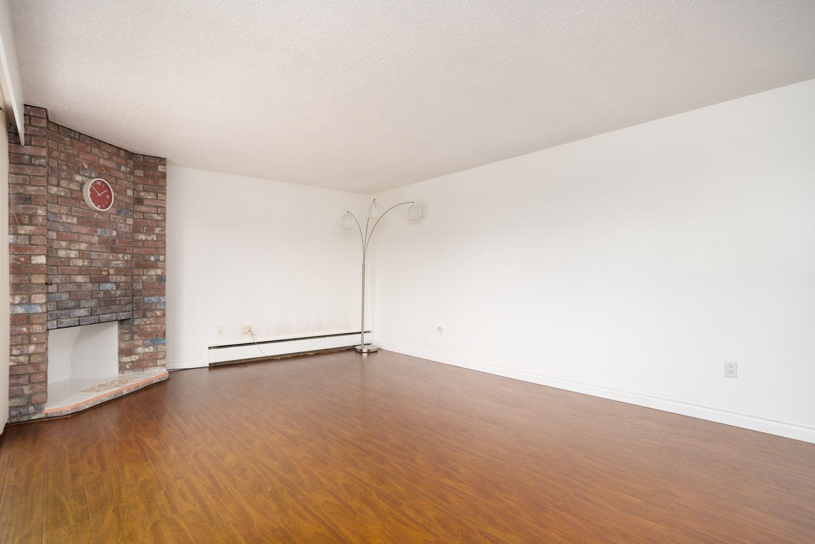 Empty room with white walls, wood floors, a corner brick fireplace, and a tall floor lamp with three lights.