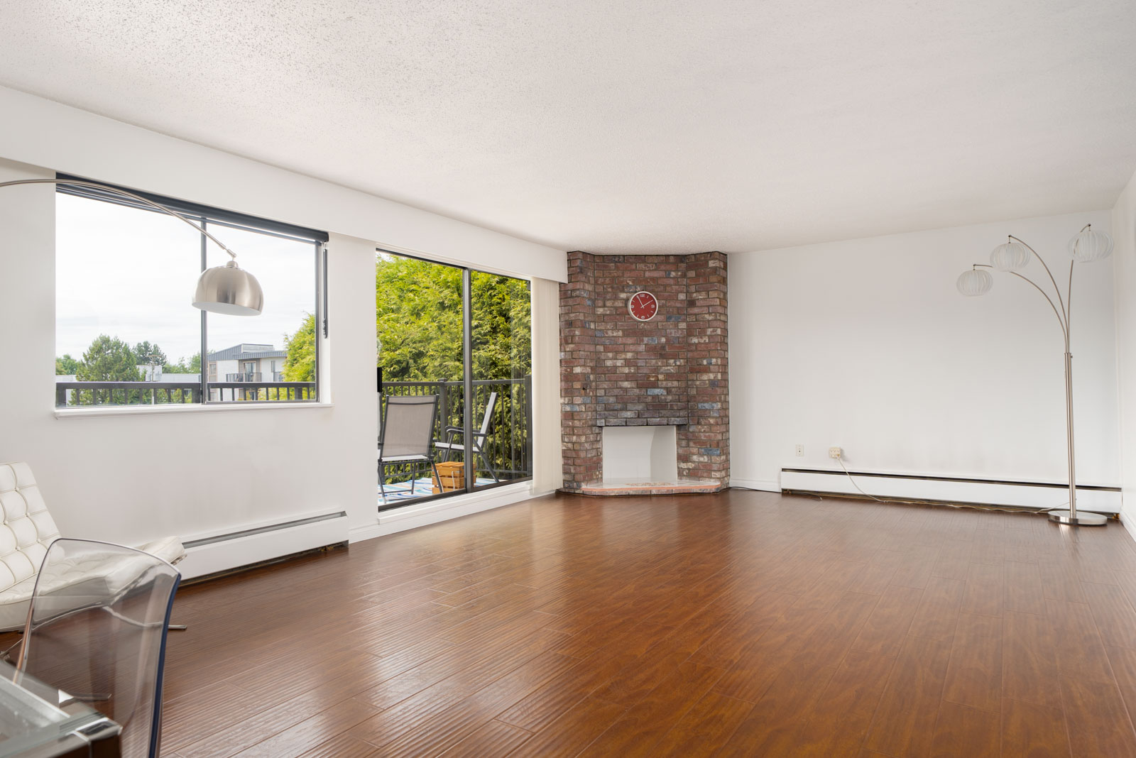 Living room with wood floors, brick fireplace, floor lamp, large windows, and sliding glass door to a balcony overlooking greenery.
