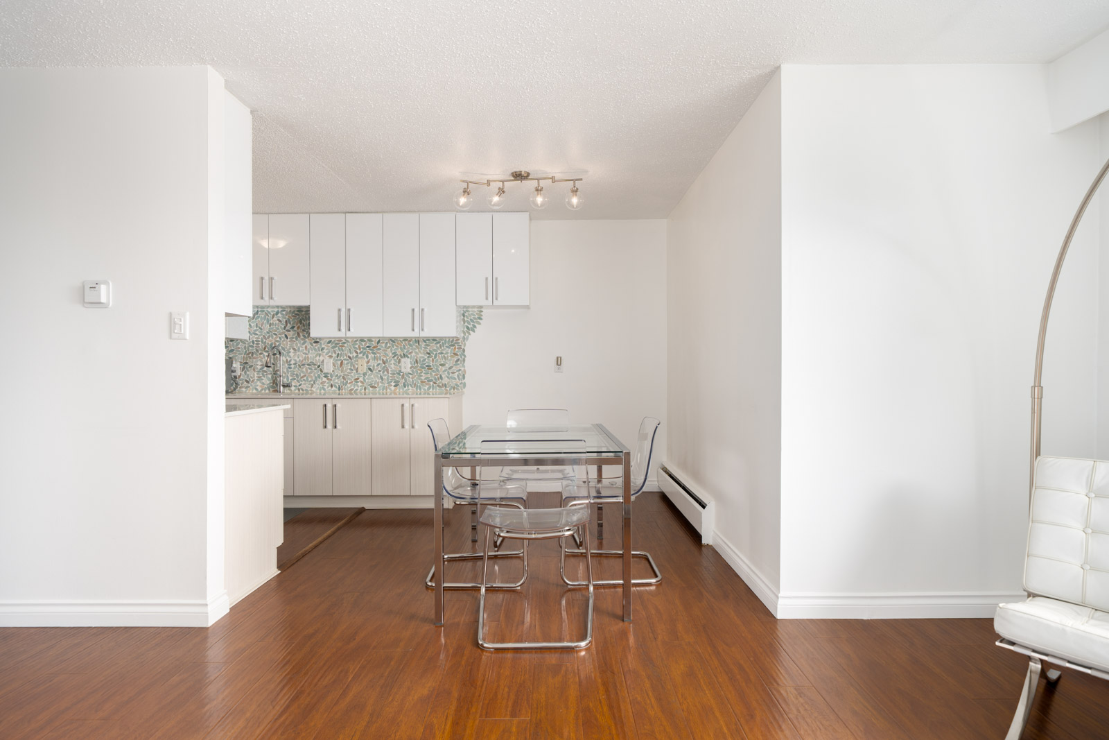 A modern dining area with a glass table and four clear chairs next to a white kitchen with patterned backsplash and wood floors.