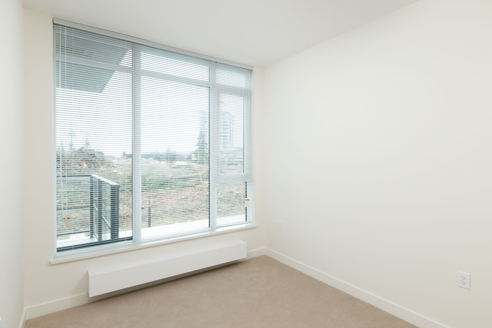 Empty room with beige carpet, white walls, large window with blinds, view of trees and buildings at 8850 University Cres, Burnaby.