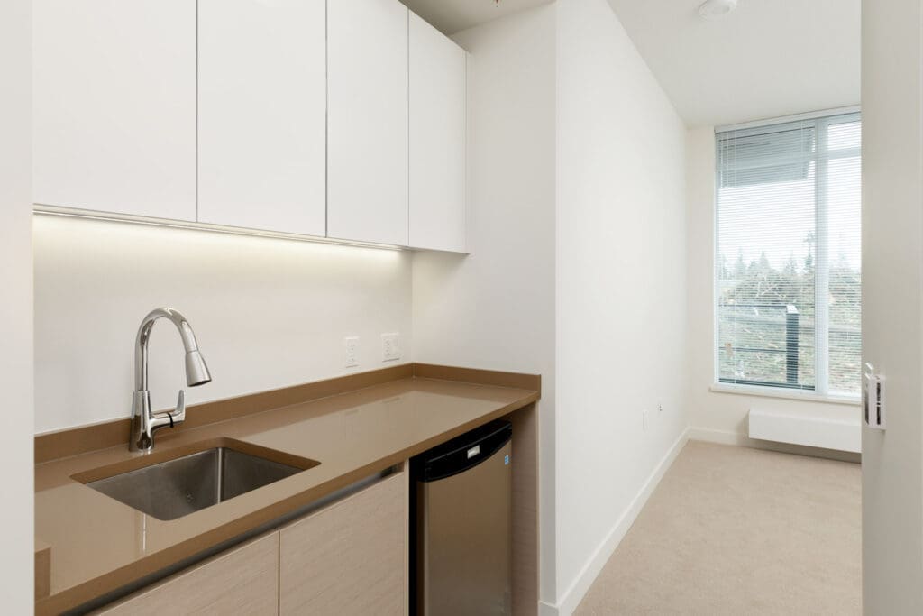 Modern kitchenette at 8850 University Cres with brown countertop, stainless sink, mini fridge, white cabinets, and neutral walls; opens to carpeted room with large window and balcony in Burnaby.
