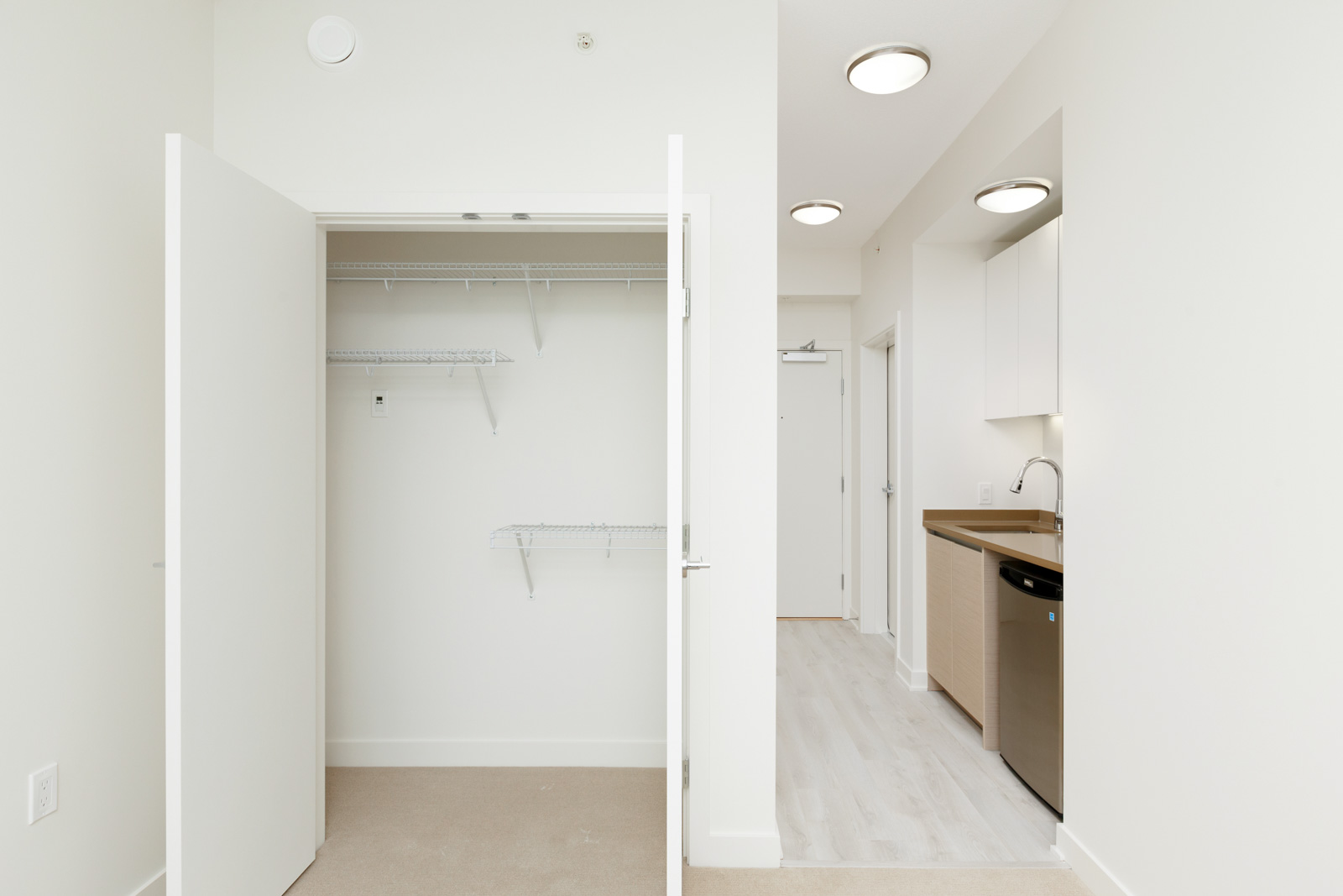 Open closet with white shelves beside a hallway to a kitchen with light wood cabinets, white walls, and ceiling lights in Burnaby.