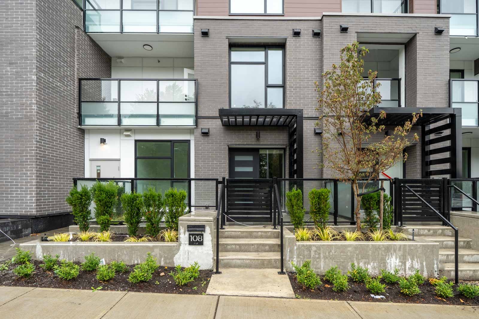 Modern townhouse exterior with large windows, a black-framed glass door, small front garden, and steps leading to the entrance. The unit number 108 is displayed near the steps.