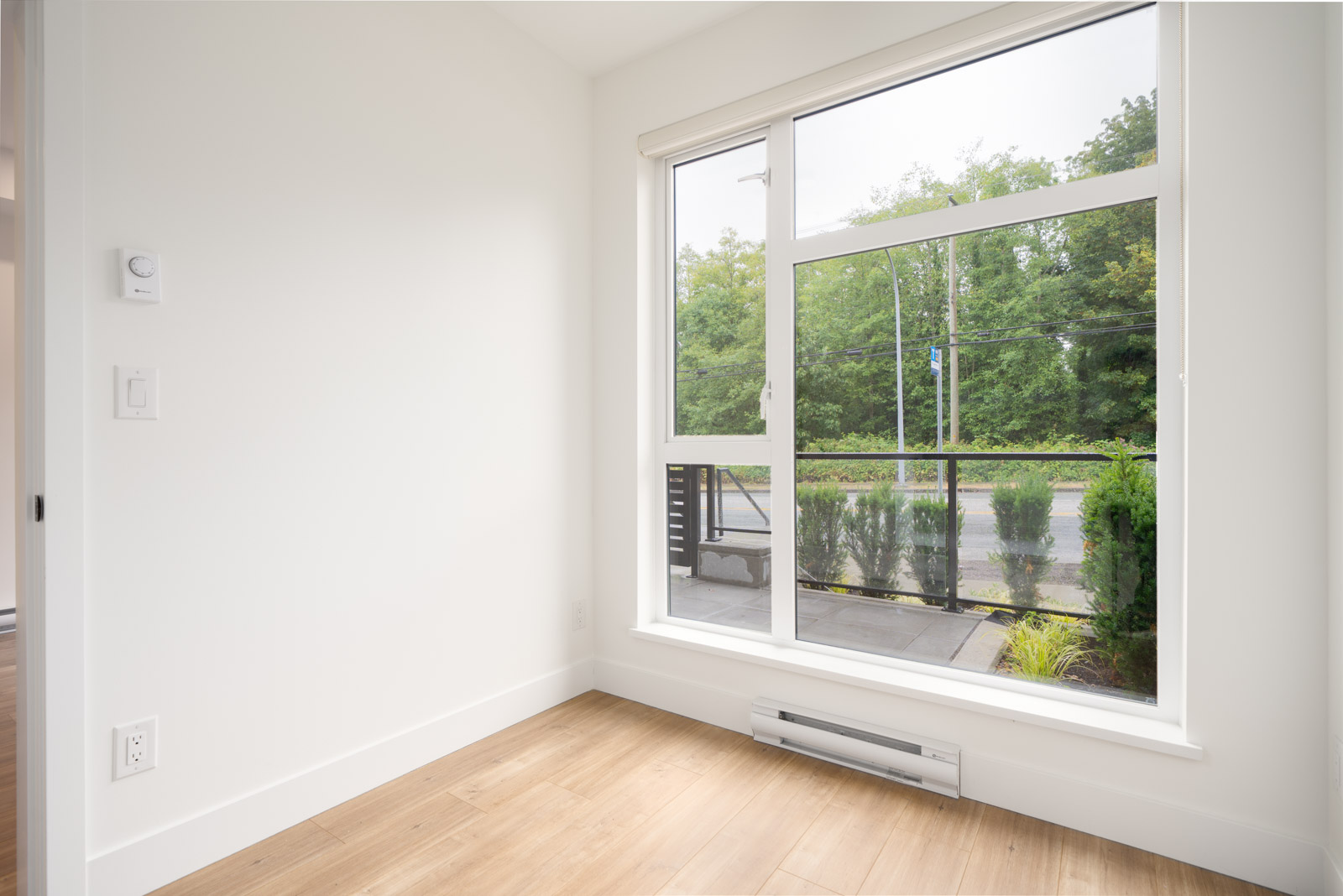 A bright, empty room with white walls, light wood flooring, and a large window overlooking a street and green trees outside.