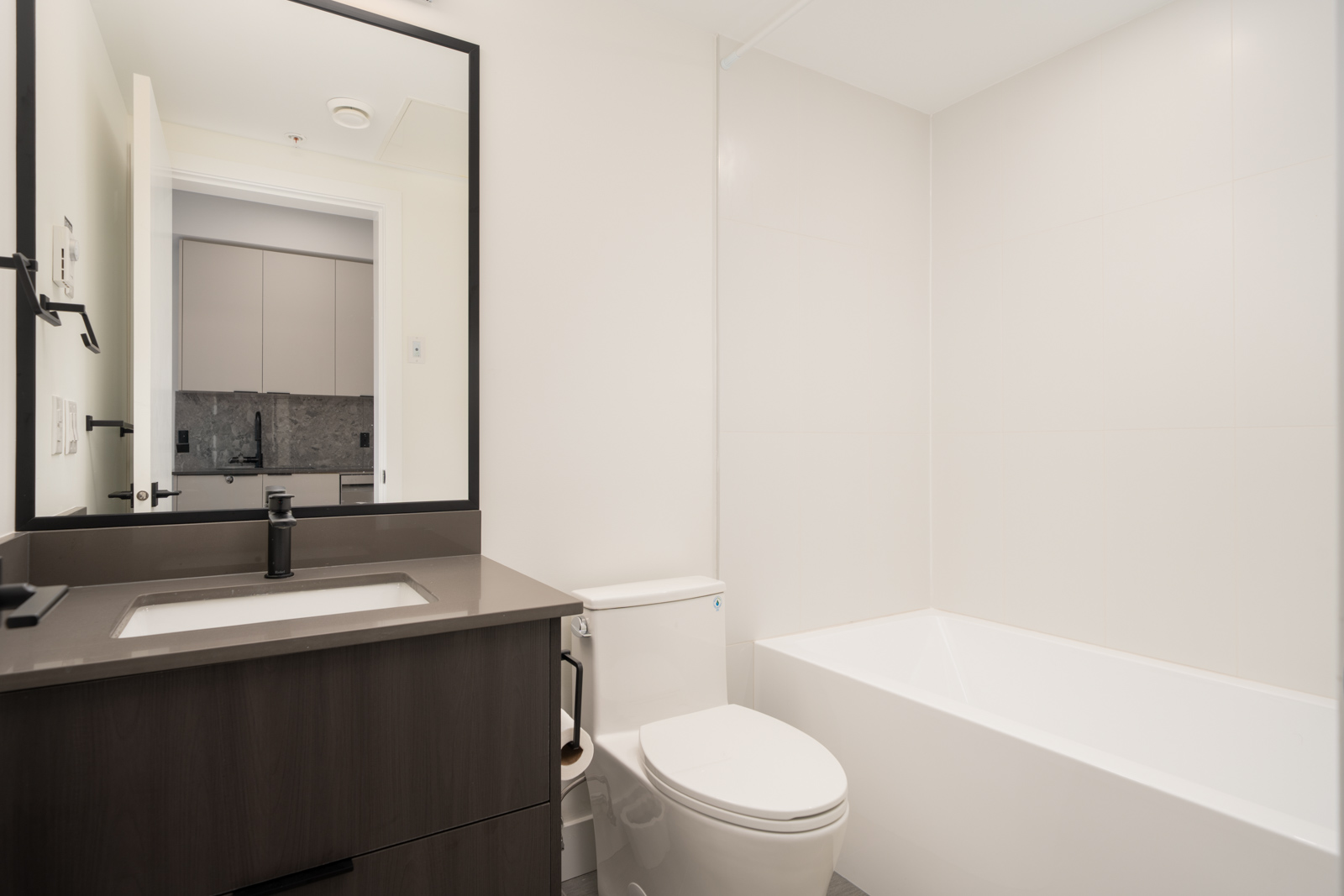 Modern bathroom with white walls, a bathtub, toilet, black vanity with sink, and large mirror above the sink. The space appears clean and minimalistic.