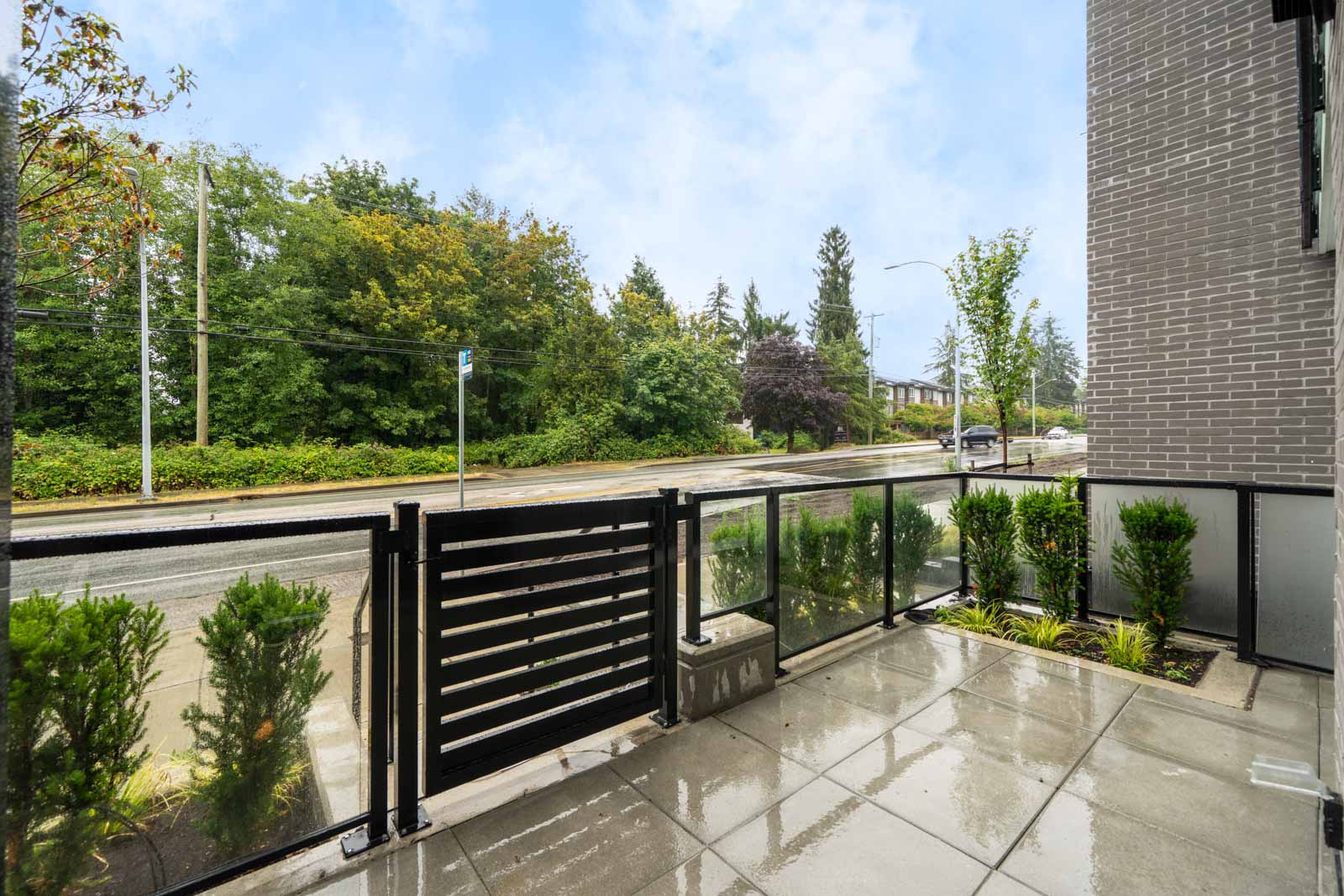 A wet patio with glass railing and gate overlooks a street lined with trees and bushes on a cloudy day.