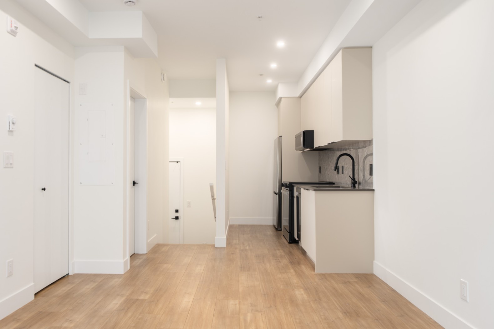 A modern, unfurnished apartment interior with wood flooring, white walls, a small kitchen area, and a hallway leading to other rooms.