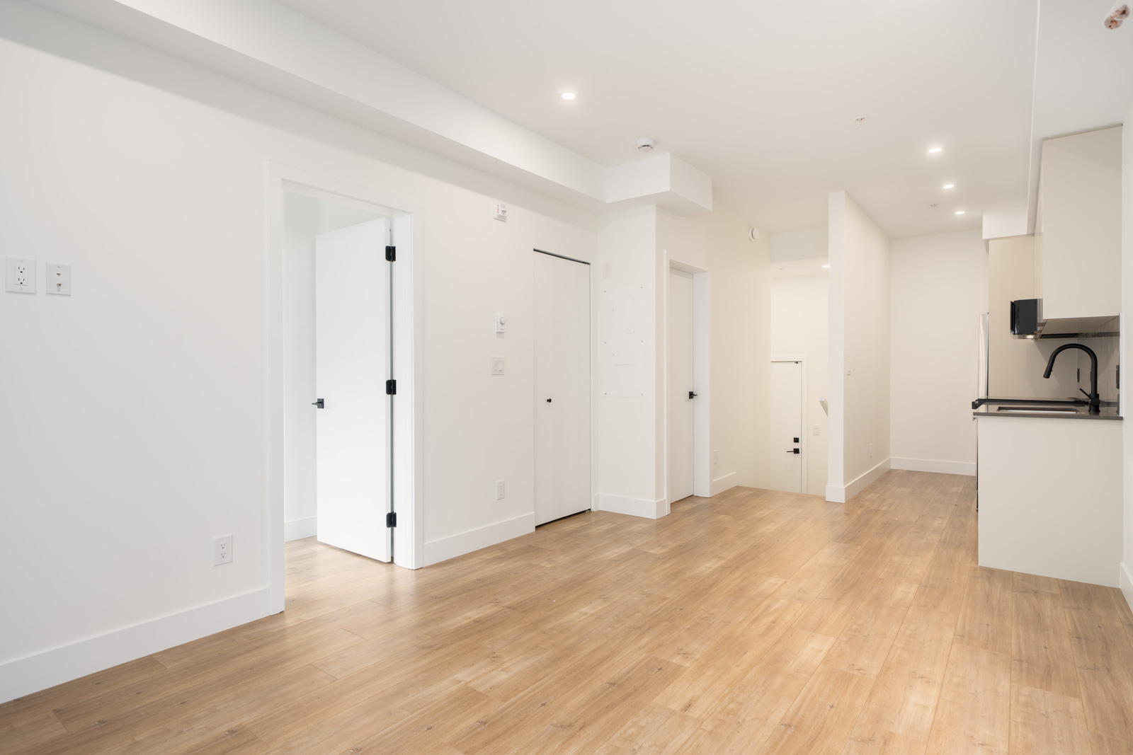 Bright, empty apartment interior with white walls, wood flooring, a kitchen area on the right, and several doors leading to other rooms.