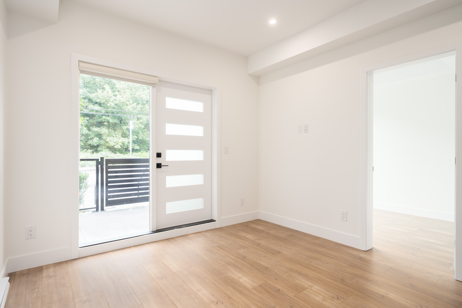 A bright, empty room with light wood flooring, white walls, a modern white front door with glass panels, and a sliding glass door leading to a balcony.