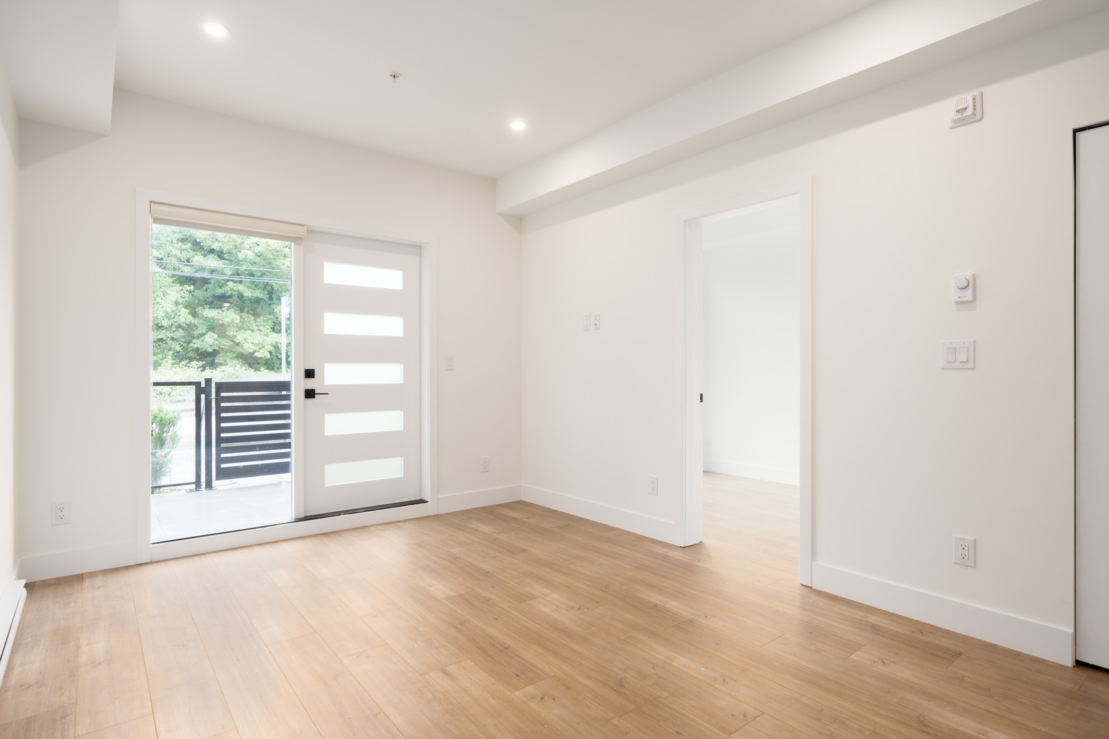 Bright, empty room with light wood flooring, white walls, a modern glass panel door leading outside, and an open doorway to another room.